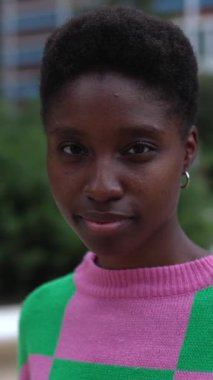 Şehrin arka planında kameraya gülümseyen modaya uygun genç Afrikalı kadın. Açık havada dururken gülümseyen kısa saçlı mutlu bir etnik kadın portresi. Dikey HD video