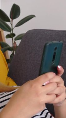 Cep telefonuna mesaj yazarken ya da sosyal medya içeriğini izlerken kanepeye uzanan mutlu genç kadının dikey HD videosu. Teknoloji yaşam tarzı ve gençlik kavramı.