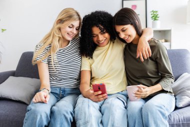 Üç tane çok ırklı genç kadın evdeki kanepede oturan akıllı telefon uygulamasında sosyal medya içeriğini izliyorlar. Teknoloji ve arkadaşlık kavramı
