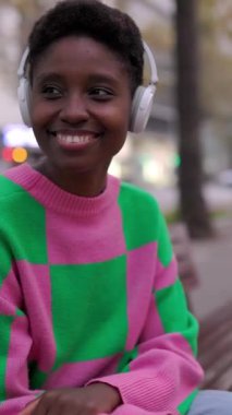Kulaklıkla müzik dinleyen Afrikalı genç bir kadın şehir bankında oturuyor. Siyahi turist kadın dışarıda dinleniyor. Seyahat ve insanlar konsepti. Dikey HD video
