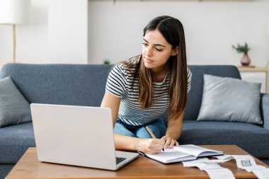 Dizüstü bilgisayarda çalışan genç bir kadın. Kanepede oturmuş ev işleriyle uğraşıyor..