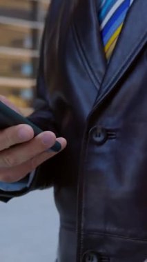 Şehirde yürürken akıllı telefon kullanan işadamının yakın görüntüsü. Uygulamada sosyal medya içeriğini kaydırırken çevrimiçi mesaj atmak, cep telefonu ekranına dokunmak ve kaydırmak. Dikey video
