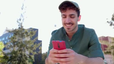 Mutlu genç adam şehir caddesinde cep telefonu kullanırken eğleniyor. Neşeli hippi General Z dışarıda günbatımı arka planında akıllı telefon uygulamasında sohbet ediyor. Teknoloji yaşam tarzı ve gençlik kavramı.