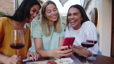 Barda aperatiflerin tadını çıkarırken cep telefonu kullanan üç genç kadın. Milenyum çeşitliliğindeki İtalyan kızları kırmızı şarap içerken akıllı telefondan sohbet ediyorlar.