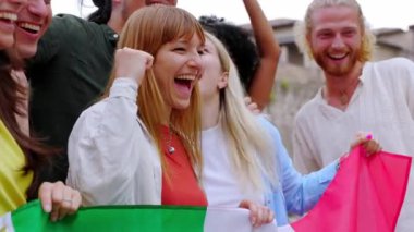 İtalyan bayrağını açık havada tutarken çığlık atan ve kutlama yapan bir grup genç. Farklı milletlerden İtalyan vatandaşlar birlik gösteriyor.