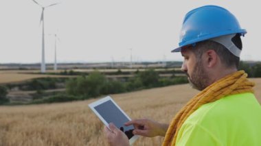 Young technician worker man with blue safety helmet and climbing equipment working with digital tablet on wind turbine farm - Renewable energy concept