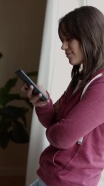 Evde rahat bir kadının cep telefonu kullandığı dikey HD video. Genç bir kadın sosyal medya ağlarından akıllı telefondan yeni içerikleri kontrol ediyor. Teknoloji konsepti. 