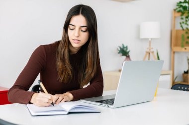 Evde dizüstü bilgisayarla çalışırken deftere yazan genç bir iş kadını. Eğitim ve e-öğrenim kavramı.