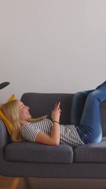 Evdeki kanepede uzanırken cep telefonu kullanan mutlu, beyaz bir kadının tam açısı. Dikey HD video. Uzayı metin için kopyala. Ekran görünümü.