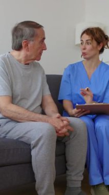 Mavi üniformalı genç kadın pratisyen hekimin evde sağlık kontrolü sırasında engelli yaşlı hastayla konuştuğu dikey HD videosu. Sağlık ve yaşlılar yaşam tarzı kavramı.