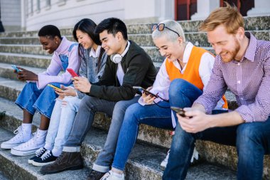 Dışarıda oturan akıllı telefonları kullanan çok ırklı genç arkadaşlar. Milenyum çeşitliliğindeki yetişkin insanlar cep telefonlarına bağımlılar ve sosyal medya içeriğini izliyorlar.