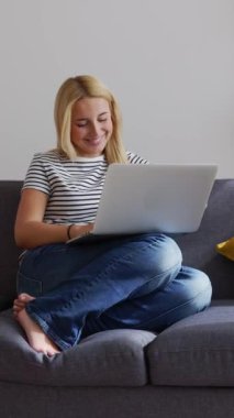 Oturma odasındaki kanepede otururken dizüstü bilgisayarda çalışan genç bir kadın. Milenyum güzeli öğrenci kız çevrimiçi çalışıyor. Dikey HD 9: 16 videosu.