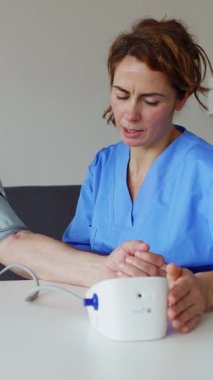 Dikey video. Evde oturma odasındaki masada oturan yaşlı adama atardamar tansiyonunu ölçen genç bir kadın sağlık görevlisi. Yaşlılık ve tıbbi yardım kavramlarında yüksek tansiyon.