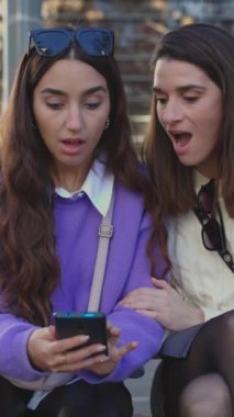 Heyecanlı genç bayan arkadaşların cep telefonundan beklenmedik mesajlar okuduğu dikey bir video. Şehir caddesinde bankta oturuyorlar. Bin yıllık gençlerin cep telefonu uygulamasından iyi haberler almasına şaşırdım.