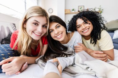 Pijamalı üç mutlu genç kız evde pijama partisi sonrası şiltede uzanmış selfie çekiyorlar.
