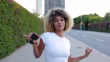 Şehir caddesinde dans eden mutlu Afro-Amerikan kadın. Binlerce yıllık etnik kızın kameraya doğru giderken telefonda dans müziğinin tadını çıkarmasını ön planda görmek. Tasasız ve mutluluk konsepti