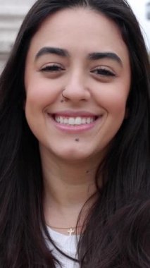 Dikey video. Dışarıda kameraya gülümseyen mutlu genç kadının portresini yakından çek. Güzel esmer kadının neşeli portresi, dişlek bir gülümseme sergiliyor..