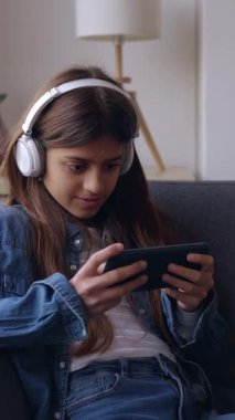 Telefon uygulaması kullanarak kulaklık takan genç bir kızın dikey videosu. Konsantre genç bayan oyuncu evdeki kanepede dinlenirken akıllı telefon kullanıyor. Boş zaman konsepti.