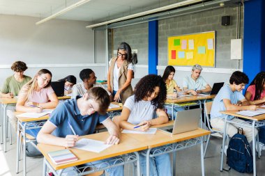 Lisede akademik muayene yapan bir grup genç. Orta yaşlı bayan öğretmen Afrikalı öğrencilere sınıfta yardım ediyor. Eğitim ve okul konseptine dönüş.