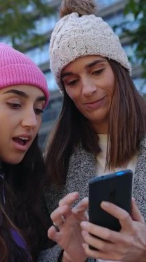 İki yetişkin kadının cep telefonuna bakıp sosyal medya haberlerinin şehir caddesinde yan yana durduğunu tartışırken çekilmiş dikey HD görüntüleri. Teknoloji ve medya kavramı.