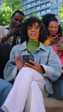 Şehir caddesinde cep telefonu kullanarak eğlenen genç bir grup insanın dikey HD videosu. Gülümseyen çok ırklı arkadaşlar dışarıda oturmuş cep telefonu uygulamasında sosyal medya içeriğini izliyorlar..