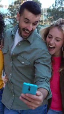 Dışarıdaki cep telefonu ekranına bakarak eğlenen genç arkadaşların dikey HD videosu. Çok ırklı insanlar birlikte gülüyor akıllı telefon ağı uygulamasında komik sosyal medya içeriklerini izliyorlar.