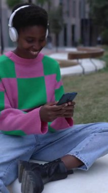 Açık havada dinlenen genç trend siyahi kadın akıllı telefonunun dikey HD videosu. Milenyum kadınları şehir bankında otururken arkadaşlarıyla cep telefonu uygulamasında sohbet ediyorlar..