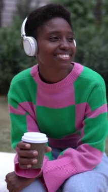 Genç Afrikalı bir kadının kablosuz kulaklık kullanarak şehir parkında oturmuş müzik dinlerken çekilmiş dikey HD videosu. Milenyum siyahi dişisi şehir molasının tadını çıkarıyor..