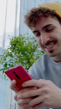 Dikey HD video. Açık havada cep telefonu kullanan genç üniversiteli hippi öğrencinin düşük açılı görüntüsü. Teknoloji yaşam tarzı ve sosyal medya kavramı.