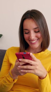 Oturma odasındaki koltukta oturan akıllı telefon cihazını kullanan mutlu ve gülümseyen genç bir kadın. Milenyum dişisi evde cep telefonu uygulamasında sohbet ediyor. Sosyal medya ve teknoloji kavramı.