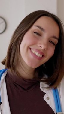 Dikey HD görüntülü gülümseyen genç bayan doktor muayene odasında duruyor. Hastane çalışanı beyaz önlüklü, klinik çalışma masasındaki kameraya bakıyor. Sağlık hizmetleri insanları kavramı