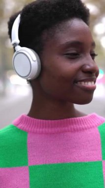 Kulaklıklı genç Afrikalı kadının dikey HD videosu. Şehir caddesinde dikiliyor. Güler yüzlü zenci kadın nesil z hipster kentsel arka planda.