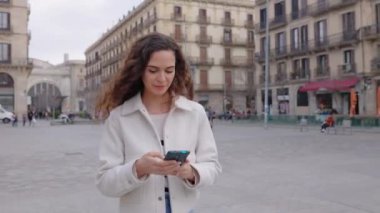 Neşeli, kıvırcık, günlük giysiler içinde, şehir caddesinde yürürken cep telefonu kullanan güzel bir kadın. Teknoloji ve şehir yaşam tarzı kavramı