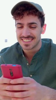 Şehir caddesinde cep telefonu kullanırken eğlenen mutlu genç adamın dikey HD videosu. Neşeli hippi General Z dışarıda akıllı telefon uygulamasında sohbet ediyor. Teknoloji yaşam tarzı ve gençlik kavramı.