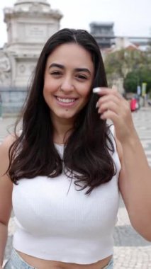 Dikey video. Lizbon, Portekiz 'deki Plaza del Rossio' da kameraya gülümseyen mutlu genç kadın. Güzel esmer bir kadının neşeli portresi. 30 'lu yaşlarda. Şehirli arka planda dişlek bir gülümseme sergiliyor..