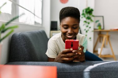 Mutlu genç bir kadın kanepeye uzanıp cep telefonu kullanıyor, evde boş vakitlerin tadını çıkarıyor, sosyal medyada geziniyor ya da arkadaşlarıyla sohbet ediyor. Teknoloji ve gençlik kavramı