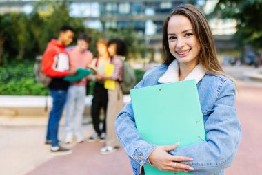 Kampüste elinde bir klasör tutan neşeli bir üniversite öğrencisinin portresi. Arka planda arkadaşları konuşuyor. Eğitim yaşam tarzı ve gençlik kavramı