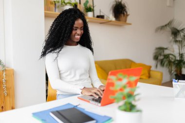 Evdeki bilgisayarda çalışan mutlu siyah kadın. Modern apartmandaki masada dizüstü bilgisayar kullanan neşeli Latin Amerikalı kadının portresi. Eğitim ve iş konsepti.