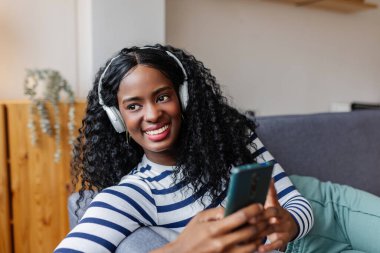 Kulaklıklı, cep telefonu kullanan, evdeki koltukta dinlenen mutlu, genç siyahi kadın. Teknoloji ve eğlence kavramı