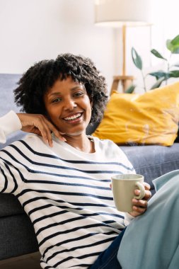 Latin Amerikalı genç bir kadının dikey portresi. Kanepede dinleniyor. Sıcak bir kış günü sıcak bir şeyler içiyor.