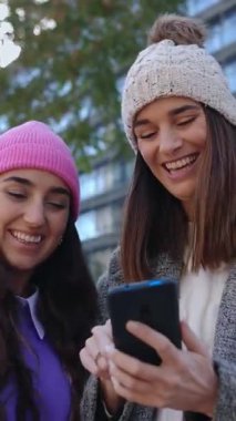 Kışlık giysiler içinde birlikte gülen ve akıllı telefon ekranına bakarken eğlenen iki güzel kadının dikey HD videosu. Milenyum kadınları telefon uygulamasında sosyal medyadan keyif alıyorlar