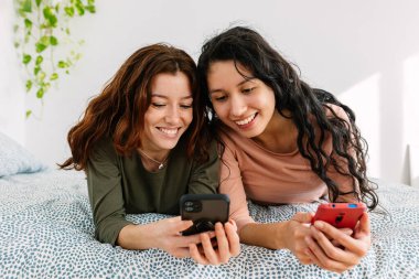 İki genç kız cep telefonlarını evde yatakta dinlenirken kullanıyor. Teknoloji ve kadın arkadaşlık yaşam tarzı konsepti.