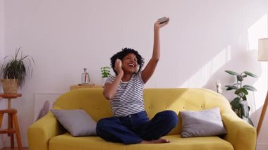 Mutlu genç bir kadın akıllı telefon uygulamasında online müzikten zevk alırken evde kanepede dinleniyor.