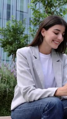 Dikey video. Şehir caddesinde cep telefonuyla konuşan genç bir iş kadını. Akıllı elbiseli profesyonel girişimci kadın dışarıda rahatlayıp akıllı telefon cihazlarını kontrol ediyor..