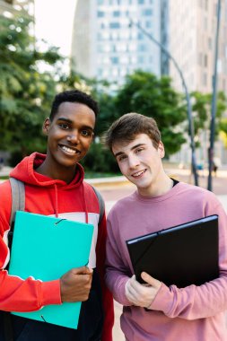 Kampüste kameraya gülümseyen iki farklı erkek üniversite öğrencisi ellerinde klasörlerle. Eğitim yaşam tarzı ve gençlik kavramı