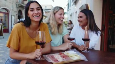 Genç, mutlu bir kadın, İtalya 'da yaz tatilinde arkadaşlarıyla birlikte dışarıda oturmuş kırmızı şarap içerken kameraya gülümsüyor. Arkadaşlık, rahatlık ve seyahat konsepti.
