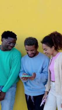 Latin Amerikalı arkadaşların sarı duvarın üzerinde cep telefonu kullanırken çekilmiş dikey HD videosu. Çeşitli bin yıllık gençler akıllı telefon uygulamasında sosyal medya içeriğini kontrol ediyorlar. Teknoloji konsepti.