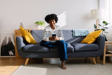 Mutlu Latin Amerikalı genç bir kızın cep telefonunu evdeki koltukta rahatça kullanması, sosyal medya içeriğinin tadını çıkarması ya da komik videolar izlemesi ön planda.