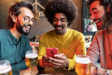 Üç erkek arkadaş bir barda birlikte vakit geçiriyorlar, bira içerken gülüyorlar ve akıllı telefona bakıyorlar.
