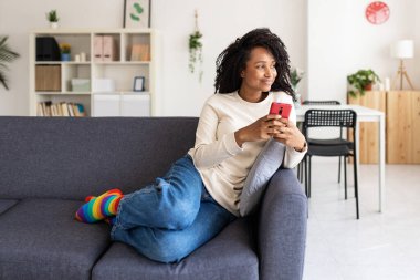 Mutlu Afrikalı genç Amerikalı kadın evinde kanepede dinlenirken elinde akıllı telefon cihazı tutuyor..